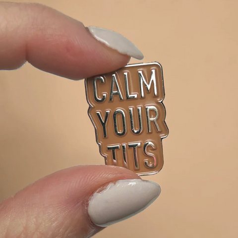 Enamel pin with 'CALM YOUR TITS' text held between fingers against a beige background