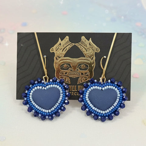 Blue heart-shaped earrings on a card with a gold design, placed on a wooden surface.