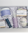 A curated Raven Song bath and body gift box filled with white crinkle paper, containing a Coastal Candles jar, Coastal Coquette spray bottle, Gardened Lavender soap bar, a dark blue tube, and a lavender bath bomb.