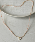 A delicate beaded necklace with small pink and gold beads and a small gold pendant, displayed on a textured white ceramic plate over a light gray surface.