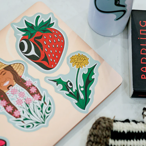 Sticker sheet with a strawberry, flower, and person design on a notebook with a mug and book in the background.