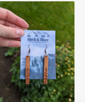 A hand holds a pair of wooden drop earrings on a branded display card outdoors. The earrings are made from light brown wood with engraved tree designs, hanging from silver fishhook wires.