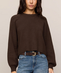 A woman wearing a dark brown knit crewneck sweater and blue jeans with a brown belt, standing against a plain light beige studio background.