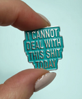 Teal enamel pin with text held by a hand against a light blue background