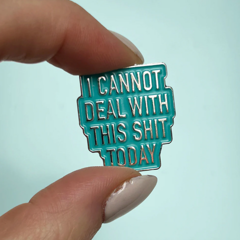 Teal enamel pin with text held by a hand against a light blue background