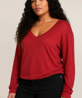 A woman with long, curly black hair wearing a red long-sleeve V-neck top and black pants, photographed against a plain light beige studio background.