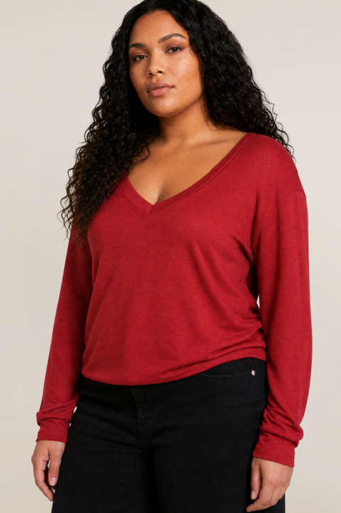 A woman with long, curly black hair wearing a red long-sleeve V-neck top and black pants, photographed against a plain light beige studio background.