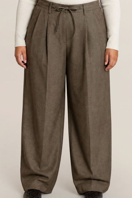 A person wearing wide-leg pleated trousers in a muted taupe-brown color, styled with a cream ribbed long-sleeve top and dark brown shoes, against a plain light beige studio background.
