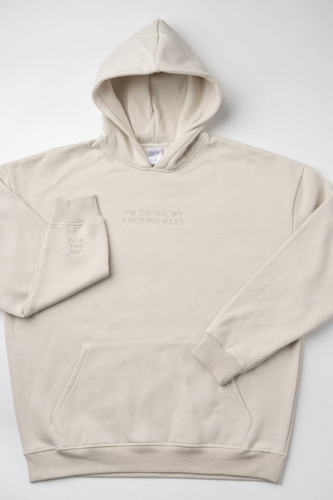 A light beige pullover hoodie with tonal embroidery on the chest reading 'I'M DOING MY FUCKING BEST' and a matching tonal sleeve graphic, laid flat on a light gray background.