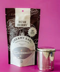 Tea package labeled 'Bush Berry Creamy Earl' with a metal tea container on a pink background