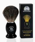 Shave brush with black handle and bristles next to its packaging on a white background