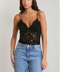A woman modeling a black lace camisole top with thin straps and blue jeans against a plain light gray studio background.