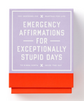 A boxed card deck titled 'Emergency Affirmations for Exceptionally Stupid Days' with a lavender top section and a bright coral-red bottom band, photographed on a clean white background.