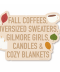 A die-cut sticker with a warm beige background featuring the text 'FALL COFFEES, OVERSIZED SWEATERS, GILMORE GIRLS, CANDLES & COZY BLANKETS' in brown lettering, decorated with small autumn-themed illustrations including leaves, a candle, and a coffee cup.