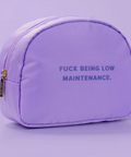 A lavender purple cosmetic pouch with a gold zipper and the text 'FUCK BEING LOW MAINTENANCE.' printed in dark purple on the front, set against a matching light purple background.