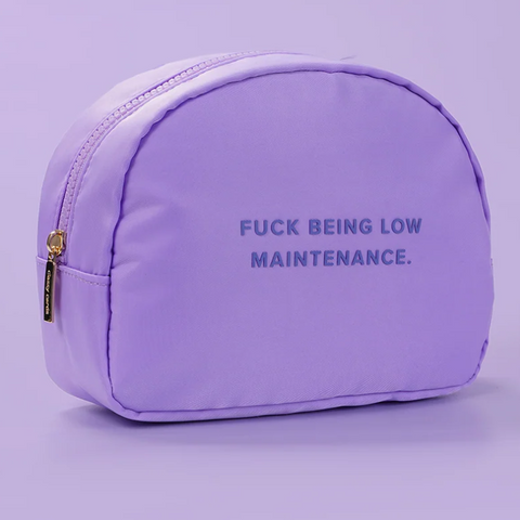 A lavender purple cosmetic pouch with a gold zipper and the text 'FUCK BEING LOW MAINTENANCE.' printed in dark purple on the front, set against a matching light purple background.