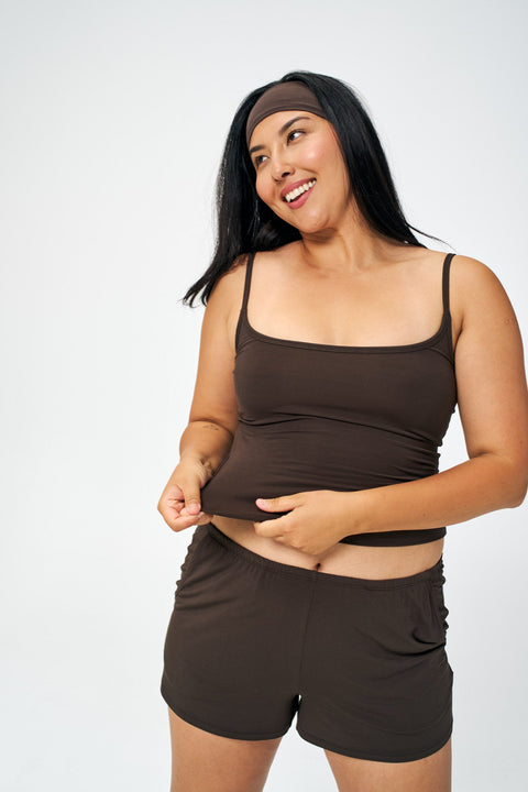 A woman wearing a matching dark brown activewear set consisting of a spaghetti-strap tank top and athletic shorts, posing against a light gray studio background.