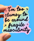 Sticker with text 'I'm too clumsy to be around fragile masculinity' held by a hand against a blue background