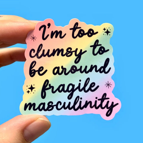 Sticker with text 'I'm too clumsy to be around fragile masculinity' held by a hand against a blue background