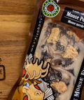 A package of Judge Fudge Factory Moose Poop Flavour, containing a clear window showing a dark brown and beige swirled mixture with crumbly beige toppings, placed on a wooden surface with the brand logo engraved on the left.