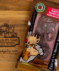 A packaged food product labeled 'Chocolate Moose Fudge Factory' and 'Raspberry Chocolate Swirl' lies on a wooden surface. The package is brown with a red top panel, cartoon moose graphics, and visible chocolate swirls inside.