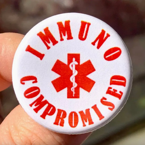 Button badge with 'IMMUNOCOMPROMISED' text and red medical symbol held by a hand.