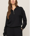 A woman modeling a black long-sleeve quarter-zip sweatshirt with a small silver zipper pull, paired with black sweatpants, against a plain light beige background.
