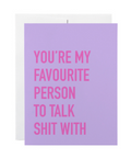 A lavender greeting card with bold pink text reading 'YOU'RE MY FAVOURITE PERSON TO TALK SHIT WITH', shown with a white envelope behind it on a white background.