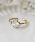 A gold-plated adjustable ring featuring a white mother-of-pearl four-petal flower charm, resting on a white textured ceramic surface with a soft-focus background.