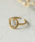 A gold-toned open ring bangle with a textured, scalloped circular edge, resting on a light marble surface with a soft-focus dried floral prop in the background.