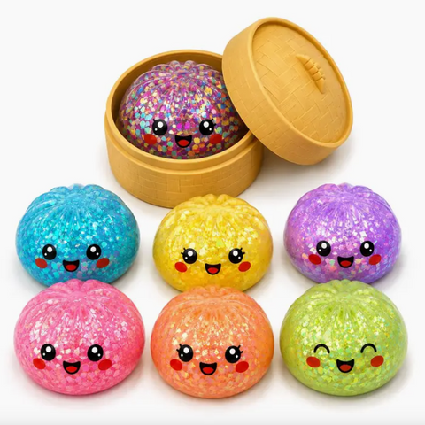 A set of seven colorful glittery squishy buns with cute cartoon faces, displayed next to a round wooden box containing one bun filled with multicolor glitter. The buns are in various colors: blue, yellow, purple, pink, orange, green, and a mix of glitter.