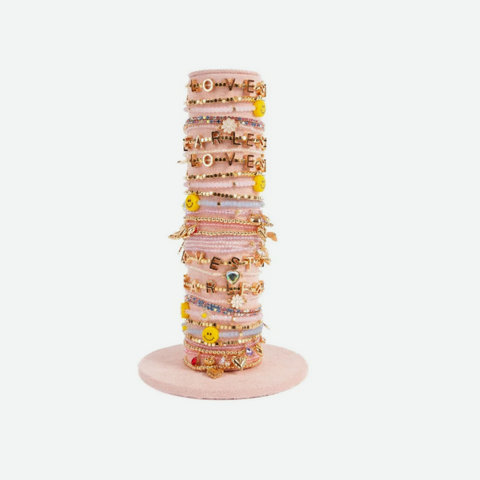 A stack of pastel pink and multicolor bracelets displayed on a matching pink stand, featuring various charms and letter beads, set against a plain light gray background.