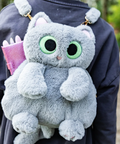 A gray plush cat backpack with large green eyes and a pink notebook tucked inside, worn on the back of a person in a dark navy jacket outdoors.