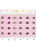 A set of six heart-shaped ring toppers displayed in a pink tray inside a white box labeled 'Great Pretenders'. The rings feature different colored heart rhinestones in purple, blue, and clear, each mounted on a gold band.