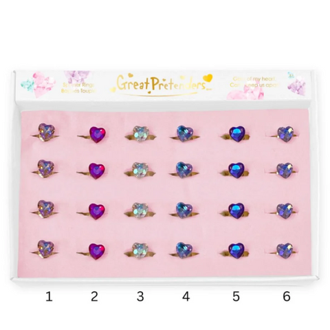 A set of six heart-shaped ring toppers displayed in a pink tray inside a white box labeled 'Great Pretenders'. The rings feature different colored heart rhinestones in purple, blue, and clear, each mounted on a gold band.