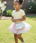 A young girl with her hair in two buns stands on a green lawn, smiling and holding a small crossbody bag with a rainbow design. She is wearing a pale yellow short-sleeve top and a sparkly pink tutu skirt.