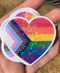 Heart-shaped rainbow sticker with glitter held in a hand against a natural background