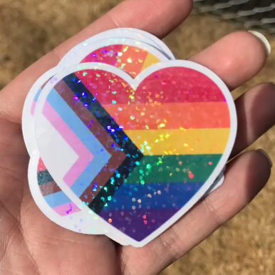 Heart-shaped rainbow sticker with glitter held in a hand against a natural background