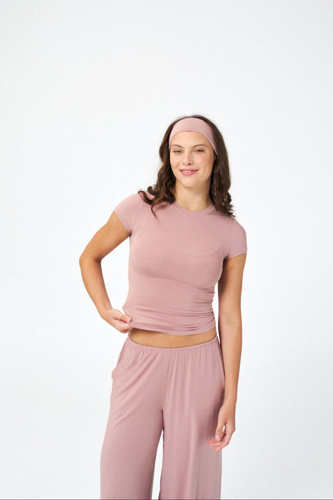 A woman modeling a matching dusty pink activewear set consisting of a short-sleeve crop top, matching pants, and a matching headband, set against a plain white studio background.