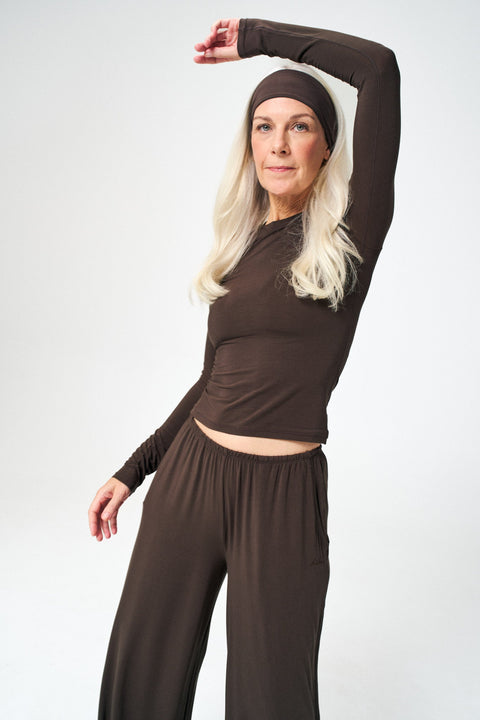A woman modeling a matching dark brown activewear set consisting of a long-sleeve cropped top, matching headband, and loose-fitting pants, posed against a light gray studio background.