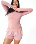 A woman posing in a matching dusty pink long-sleeve top and shorts set against a solid white background.