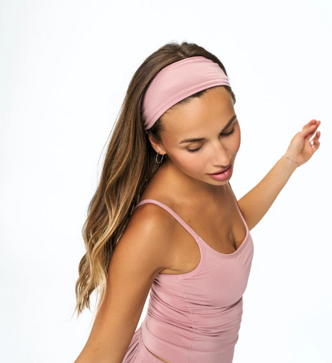 A woman wearing a dusty pink athletic headband and matching pink sports bra, posing against a solid white background.
