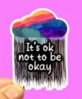 Sticker with 'It's ok not to be okay' text and a colorful cloud design on a purple background.