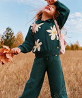 A child wearing a dark teal knit matching set with large white daisy flowers, standing in a dry grass field under a blue sky. The outfit includes a long-sleeve top and wide-leg pants, and the child is holding autumn leaves.
