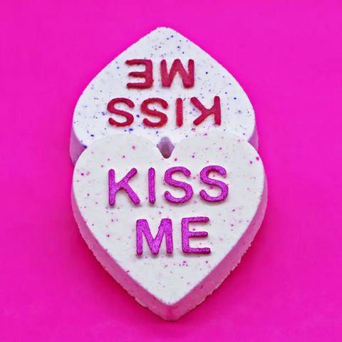 Two heart-shaped candy pieces with 'WE KISS ME' text on a pink background