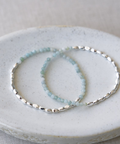 Two beaded bracelets, one with light blue-green round beads and one with silver faceted beads, displayed on a round white ceramic dish.