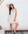 Woman in a white dress standing against a white wall with palm leaves above her.