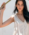 Woman in a white sleeveless top standing against a light background with a palm leaf.