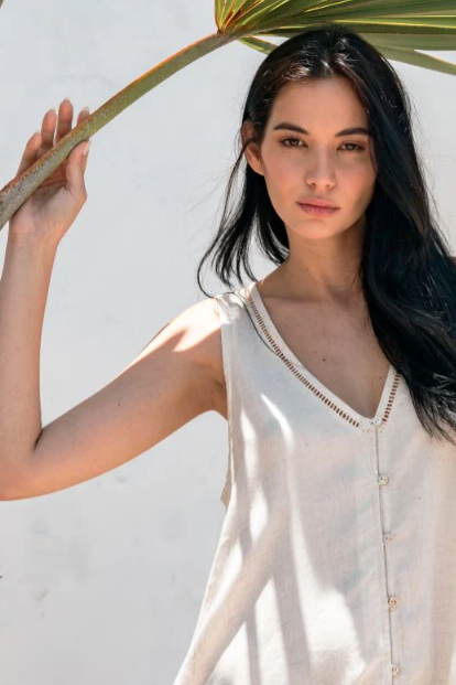 Woman in a white sleeveless top standing against a light background with a palm leaf.