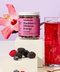 A jar of lake&oaks Blackberry Hibiscus Energy drink mix displayed on a white pedestal next to a glass of red hibiscus tea with ice, styled with fresh berries and a pink hibiscus flower against a lavender background.
